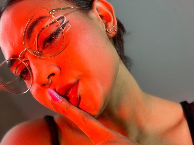 LilithRussoo with red lighting, wearing glasses, nose ring, pink nails, finger touching lips, black top, ear ring, and short dark hair, looking slightly down, serious expression.