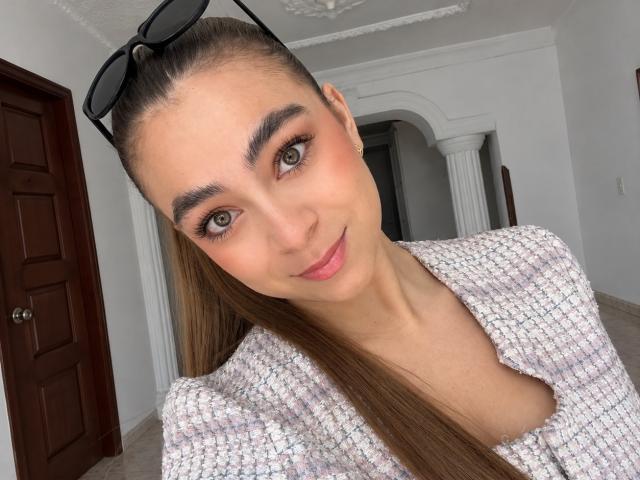 AriannaBlake with long brown hair, black sunglasses on head, wearing a white textured top, smiling at camera, brown eyes, pink lips, small nose, white wall, wooden door.