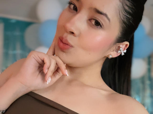 EmmaLombardi has pouty lips, a cute face, and a dark ponytail. She's touching her chin with a hand, wearing a brown top and diamond earrings.