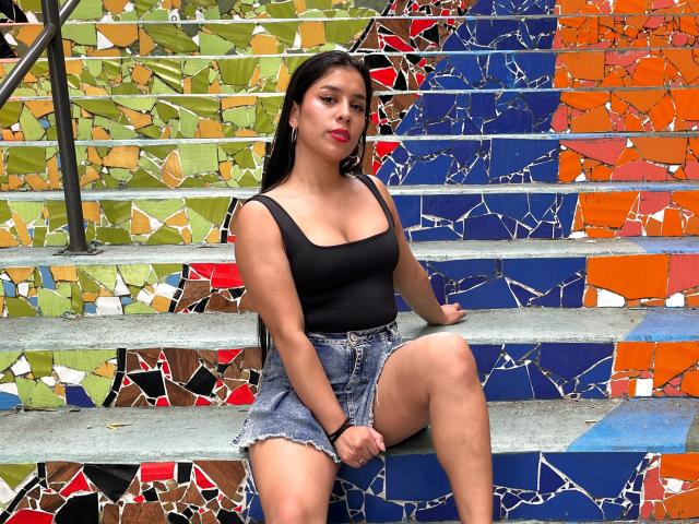 MelodyRoss with long black hair, red lipstick, black tank top, and denim shorts, sitting on colorful, mosaic steps, showing her ass and small tits.