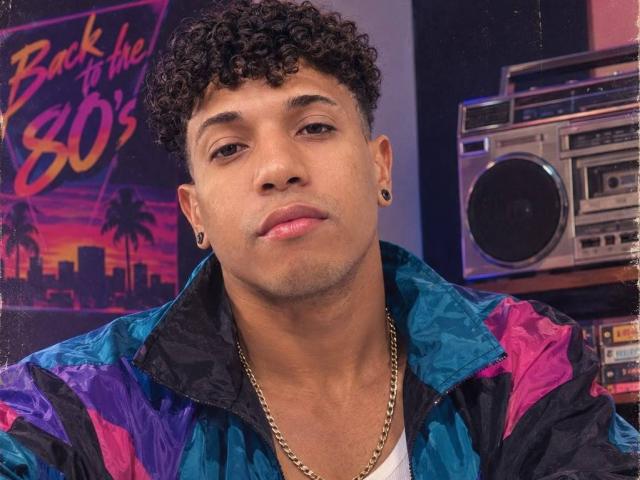 Yoximanuelprime, a curvy, dark-skinned dude with short curly hair, wearing a colorful jacket, gold chain, and black ear gauges, looking serious with a slight pout.