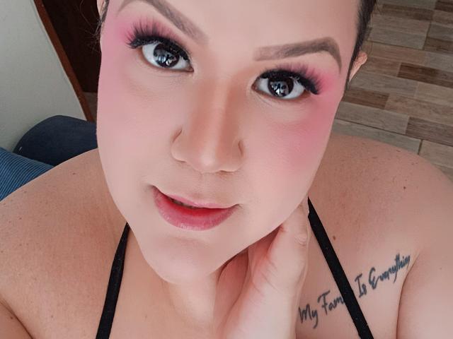 GiiannaMorrys has pink eyeshadow, long eyelashes, and a tattoo on her right shoulder. She's looking up with a slight smile, touching her neck with her right hand.