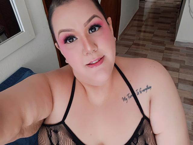 GiiannaMorrys with pink eye makeup, black mesh top, nose ring, and tattoo saying "My Time to Shine". Her tits are visible through the mesh, smiling at the camera.