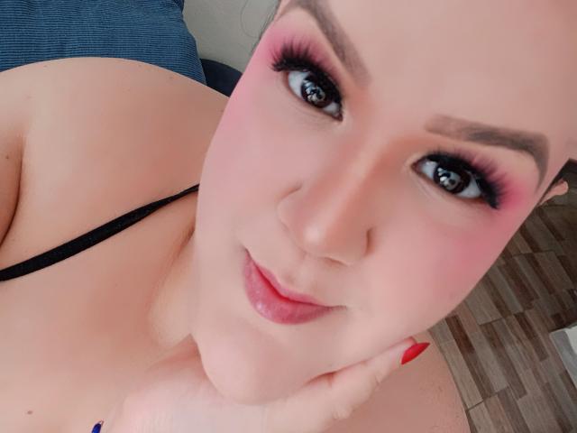 GiiannaMorrys with pink eyeshadow, black eyeliner, and red lipstick, smiling, showing bare shoulders, black strap, and red nails, holding cheek. She has fair skin and brown eyes.