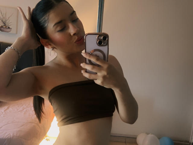 EmmaLombardi taking a selfie, pouty lips, brown strapless top, toned midriff, long black hair in a ponytail, holding a phone with a ring case, dim room.