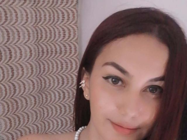 Ferlino69 has fair skin, dark brown hair, black eyeliner, small earrings, and a pearl necklace, smiling with subtle pink lips. Her eyes are green, and she looks cute.