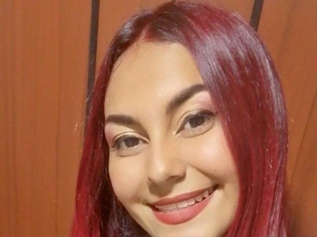 Ferlino69, met rood haar, lachend, met roze oogschaduw en roze lippenstift, heeft een schattig gezicht en laat haar tanden zien. Haar borsten zijn niet zichtbaar op deze foto..