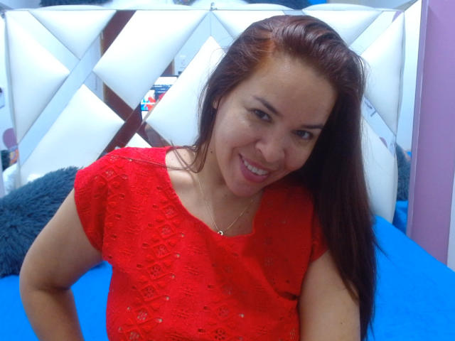 LizethBells has red lace top, medium tits, brown hair, smiling, blue bed, white diamond-patterned headboard, gray pillow, necklace, and pink wall in background.