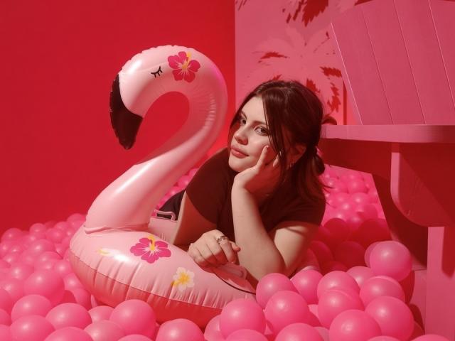 MicheMeow is laying in a pink inflatable flamingo with flower decals, surrounded by pink balloons, wearing a black top, and resting her face on her hand.