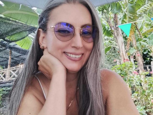 EvaNathaly with long gray hair, smiling, wearing purple-tinted aviator sunglasses, and a gold necklace, resting her chin on her hand, showing off her cute earrings.
