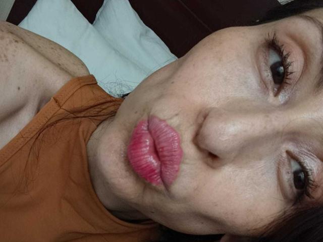 AliceRossiX has pink pouty lips, brown eyes, and light skin, wearing an orange top, with dark hair, lying down, making a pouty face.