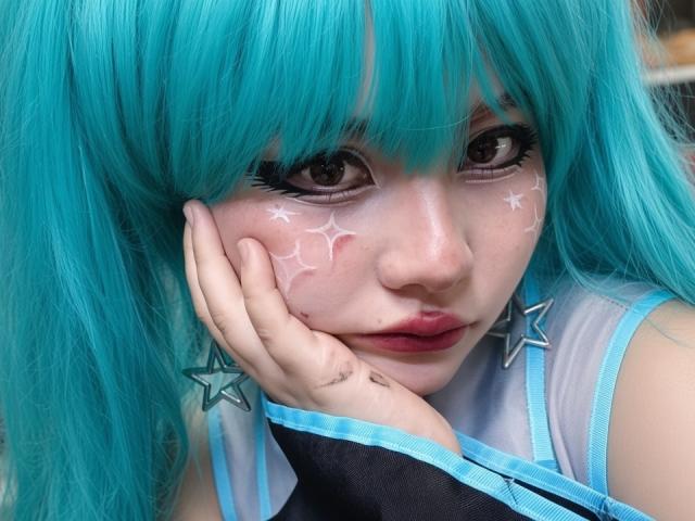 MicheMeow con i capelli color acquamarina, trucco a forma di stella bianca sulle guance, mano vicino al viso, occhi azzurri, indossa orecchini a forma di stella e una maglietta blu e nera. Le sue labbra sono leggermente dischiuse..
