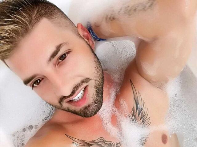TommyDum with a beard, smiling, in a bathtub of foamy water, showing tattoos on his right arm and chest, with short blonde hair and a blue necklace.