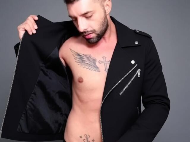 TommyDum with a black jacket open, showing a tattooed chest and abs, and a small cross tattoo below his navel, has a trimmed beard and short brown hair.
