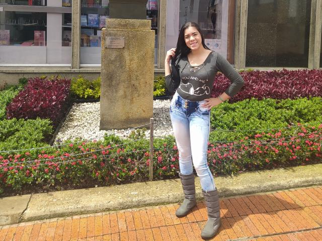 KarlyGreyKiss has dark hair, a green long-sleeve shirt with "Love" text, white and blue jeans, and gray boots, standing on a brick sidewalk with a garden.