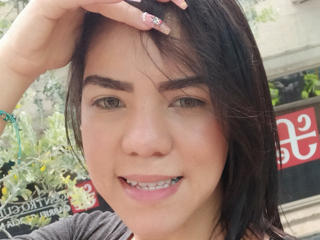 KarlyGreyKiss has dark brown hair, green eyes, and a small smile with visible teeth. She's touching her head with a hand, showing pink nail polish and a green bracelet.