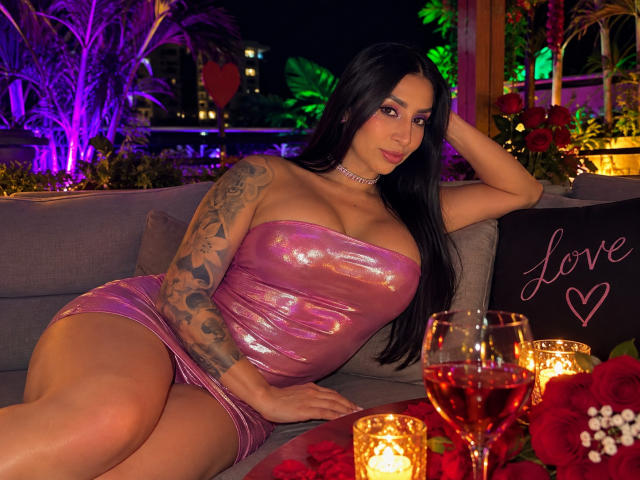 FransheskaParker with big tits, black hair, tattooed arm, wearing shiny pink dress, laying on grey couch, chandelier earrings, red wine glasses, candles, and red roses in front.