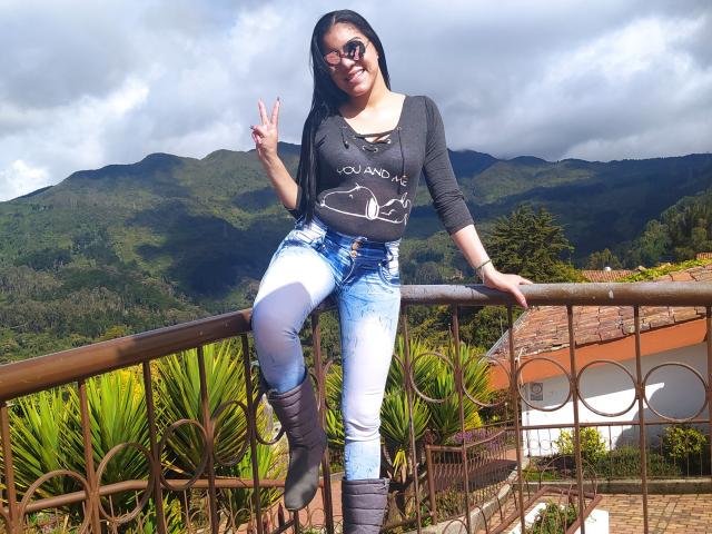 KarlyGreyKiss with dark hair, glasses, smiling, black long-sleeve, white and blue jeans, black boots, making a peace sign, standing on a balcony with mountains in the background.
