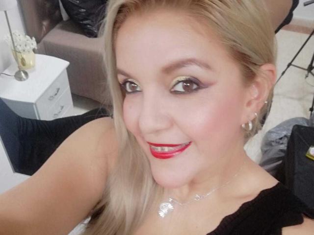 LadymorMorro, com cabelo loiro, batom vermelho e vestido preto, sorrindo para a câmera, usando brincos de argola e colar de prata, em um selfie com maquiagem suave e delineador discreto..
