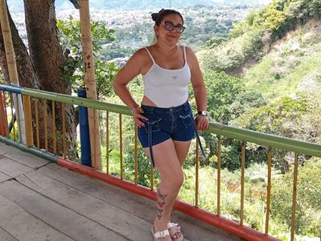 LadymorMorro con pechos medianos, camiseta blanca, pantalones cortos vaqueros azules, sandalias blancas, tatuaje en la pierna derecha, de pie en una cubierta de madera, con las manos en la barandilla, usando gafas, sonriendo..