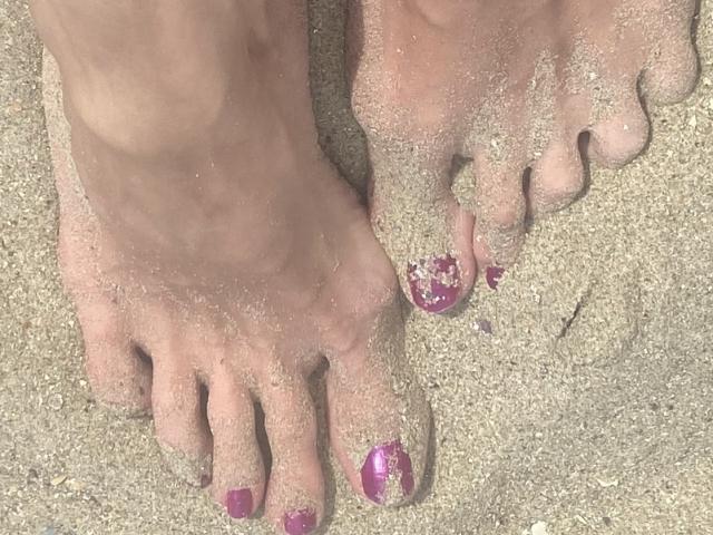 Nalini's sandy feet with pink toenails, some covered in sand, standing on a beach. Her toes are slightly apart, showing the texture of the sand on her skin.