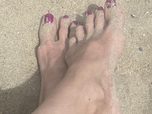 Nalini's sandy feet with purple-painted toenails, slightly dirty from the beach, are shown in a top-down view. Her toes are covered in small grains of sand.