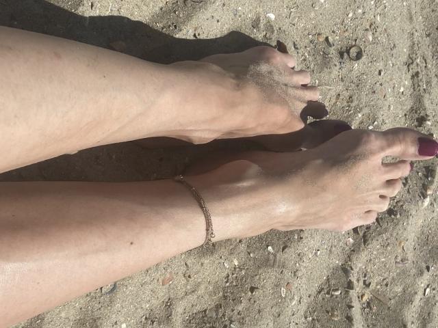 Nalini's bare feet, with sand on them and purple-painted toenails, are crossed at the ankles, wearing a thin anklet, on a dirty, sandy beach.