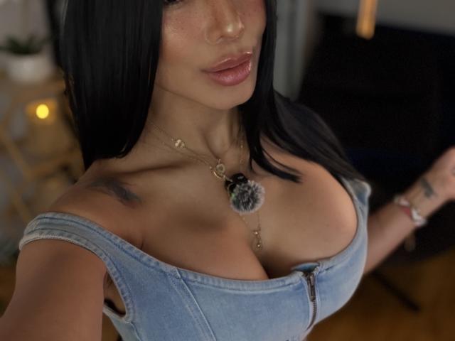 SandraXSeins avec de grandes seins, portant un débardeur en jean bleu à épaules dénudées, cheveux noirs, lèvres roses brillantes, collier en or, tatouages sur les épaules, dans une pièce éclairée doucement..