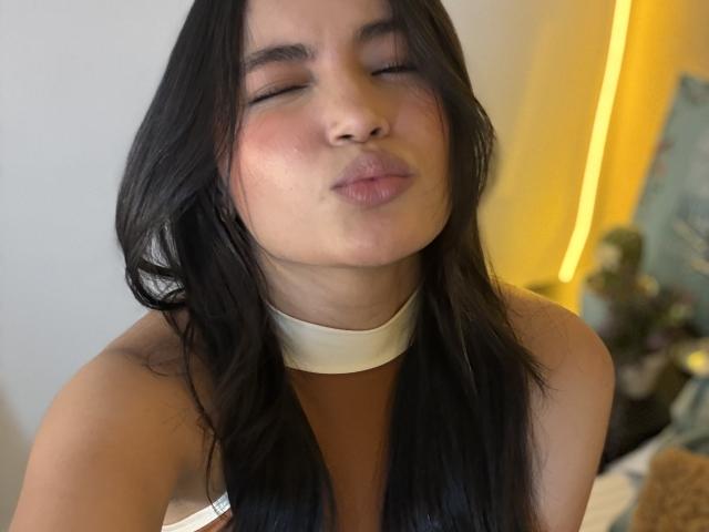 MiiaPalmer with closed eyes, puffy lips, and a light skin tone, wearing a white choker, has long black hair, and is making a kissy face. Boobs not visible.