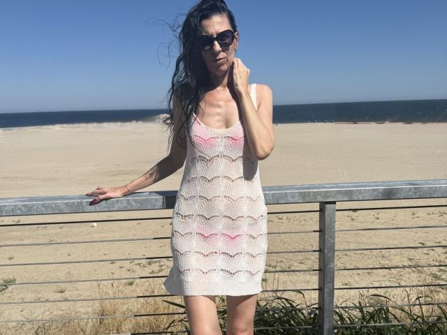Nalini está de pie en una playa, vestida con un vestido blanco y rosa de crochet, gafas de sol negras y cabello largo y ondulado, con una mano apoyada en una barandilla de metal. Está mostrando sus pechos..