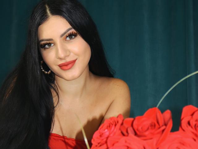 JasmineBryze with long black hair, red lipstick, and hoop earrings, smiling at red roses in the foreground, bare shoulders, dark green curtain background.