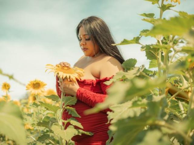 BiancaForest met donker haar, een rode jurk met een open schouder, een zonnebloem in haar hand, omringd door hoge zonnebloemen, een blauwe lucht op de achtergrond, kijkt naar beneden met een zachte uitdrukking, een zonnige dag..