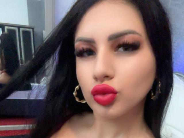 JasmineBryze with full red lips, long black hair, thick eyelashes, and gold hoop earrings, making a pouty face. Her tits are not visible.