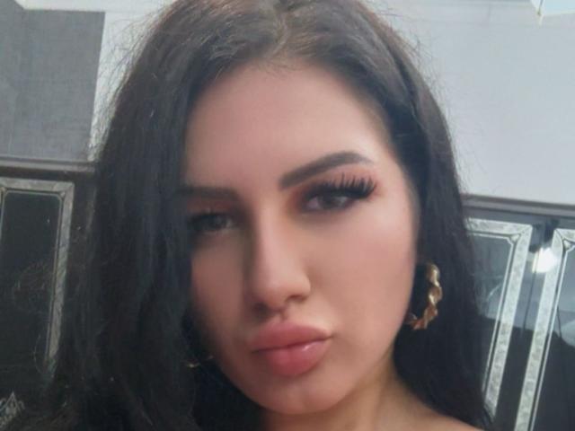 JasmineBryze has long black hair, thick eyebrows, long eyelashes, and full pink lips. She's wearing gold hoop earrings and looking slightly to the side with a serious expression.
