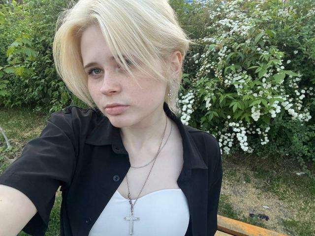 Xochitl Alejo, rubia, con el cabello peinado hacia un lado, camisa negra, top blanco, collar con una cruz, expresión de labios fruncidos, un arbusto verde con flores blancas en el fondo, sentada en un banco de madera..