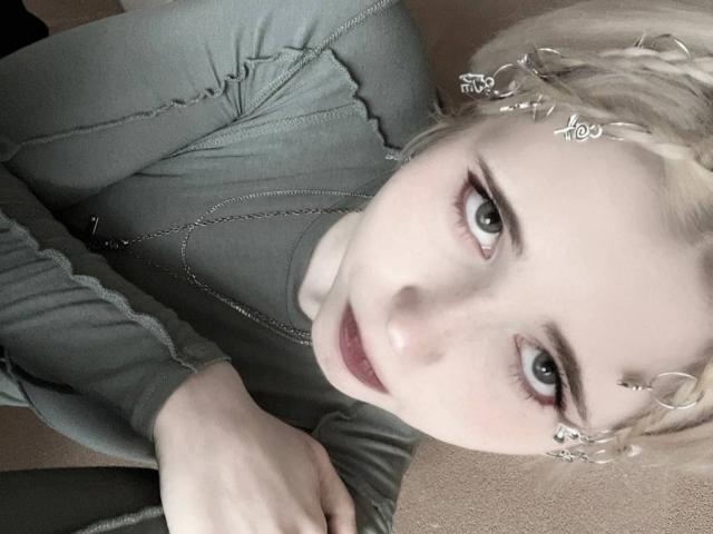 XochitlAlejo with pale skin, blonde braided hair, green eyes, and a silver headpiece, wearing a grey long-sleeve top, looking up with a slight smile.