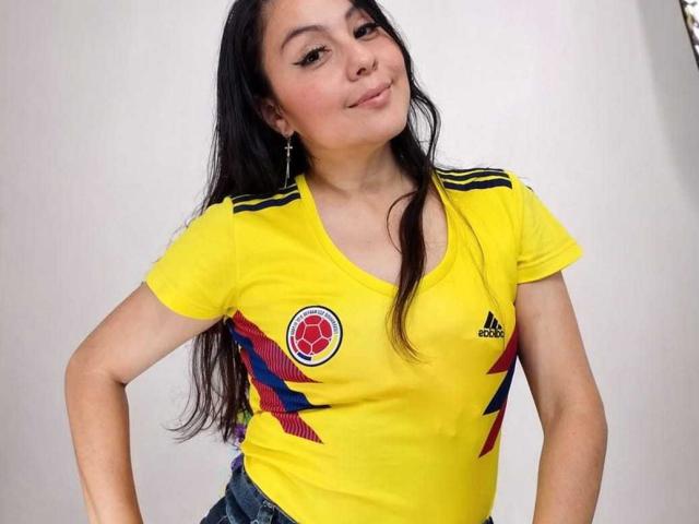 GabrielaOrozco has long black hair, a nose ring, and is wearing a yellow Colombia soccer shirt with red, blue, and black stripes, and denim jeans, smiling.