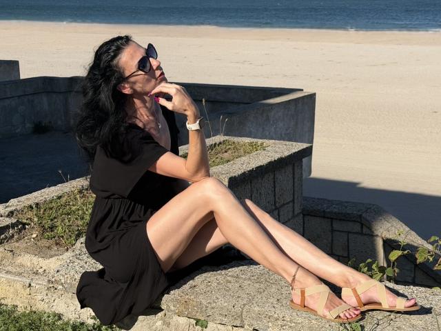 Nalini with black hair, wearing a black dress, sunglasses, and tan sandals, sitting on stone, legs crossed, holding phone to ear, beach and blue ocean in background.