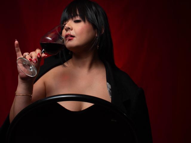 PandoraForster69 with black hair, red lipstick, and red nails, sipping red wine, holding glass to lips, black jacket, visible cleavage, black chair in front, dark red background.