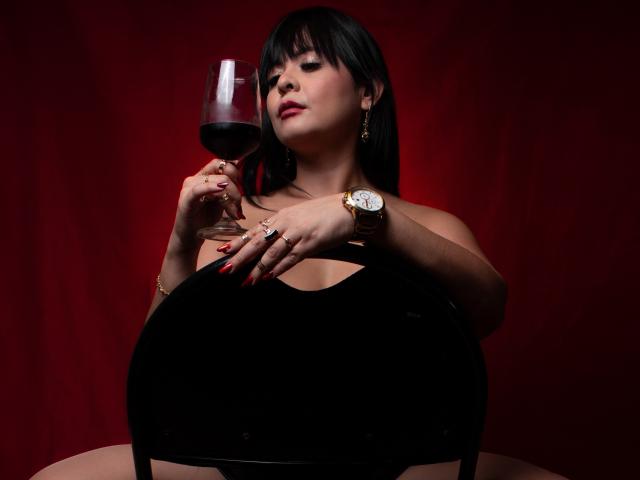 PandoraForster69 with black hair, red lipstick, and gold watch, holding glass of red wine, sitting on black chair, showing off her big tits, against red background.