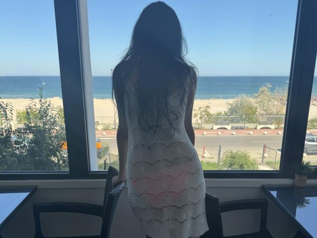 Nalini stands with her back to the window, wearing a white, lace-up dress, showing her ass. She has long, wavy dark hair and looks at a beach outside.