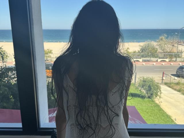 Nalini with long, wavy black hair, facing a window, wearing a white, loose-knit top, showing her back, standing in front of a beach and ocean view.