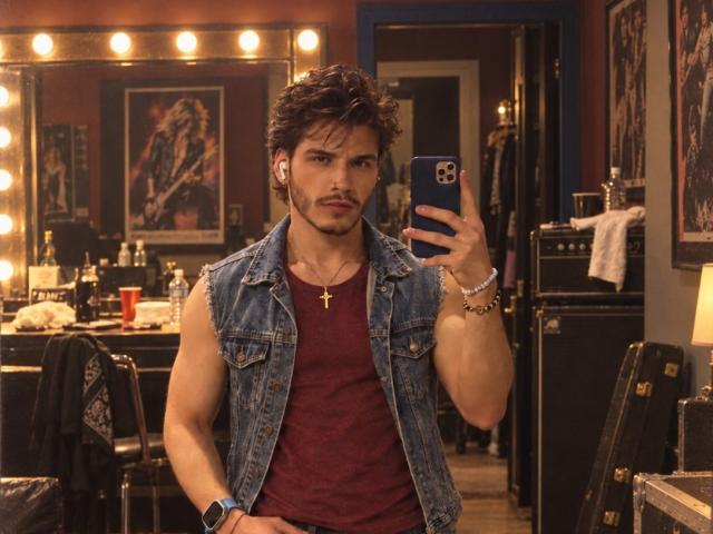 LianFerrer, musculosa, com cabelo castanho curto, vestindo uma camiseta vermelha, colete jeans, um colar com uma cruz e uma pulseira no pulso, tirando uma selfie no espelho em um quarto com pouca luz..