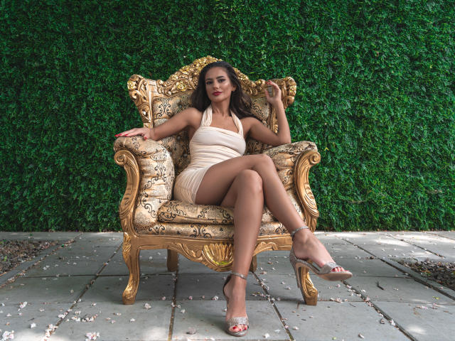 JocelynnKey with big ass, small tits, and a tan pussy, sits on an ornate chair, wearing a beige dress and silver heels, against a green wall.