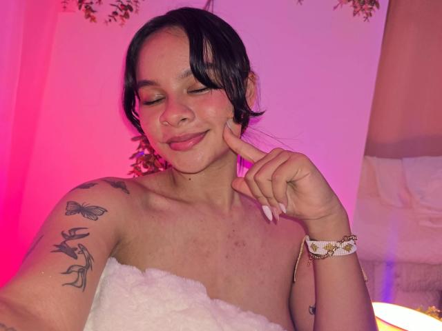 Yurimy with dark hair, closed eyes, and pink blush, touching her cheek, wearing a white strapless dress, butterfly tattoos on her arm, white bracelet, pink and purple light.