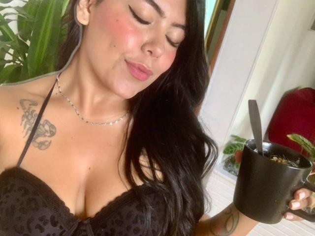 CatalinaReiyna with black lace bra, showing her big tits, holding a black cup with a spoon, long black hair, pink blush, tattoo on left shoulder, necklace, smiling.