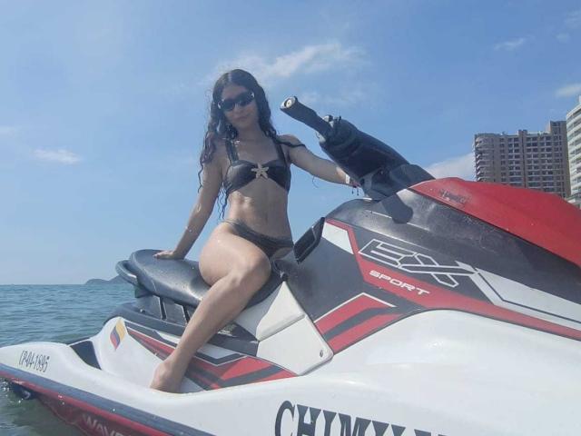 FluffydirtyMind avec un bikini noir mouillé, assise sur un jet ski rouge et blanc, ciel bleu, océan, portant des lunettes de soleil, cheveux longs et bouclés, petites seins, et un sourire sexy..