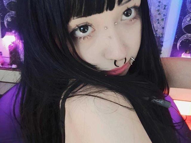 Vickmon met zwart haar, blauwe ogen, een neusring, een septum piercing en een lip piercing, kijkt over haar schouder, toont haar blote schouder, heeft bleke huid en subtiele make-up in een paars verlichte omgeving..