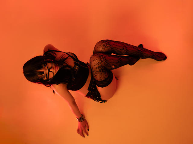 Vickmon, gekleed in zwart kanten lingerie, met stippenmotief kousen en een bril, zittend op de vloer, armen achter haar rug, benen gebogen, met een rood-oranje belichting, waarbij haar billen en borsten zichtbaar zijn..
