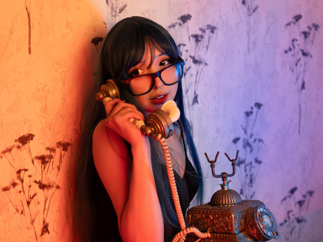 Vickmon met lang zwart haar, die een bril draagt en een ouderwetige telefoon aan haar oor houdt, ziet er schattig uit, gekleed in een rode top, en staat naast een vintage telefoon tegen een muur met een structuur..